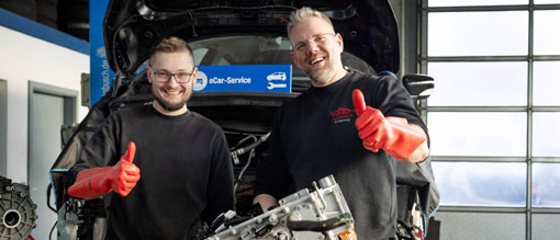 Foto: zwei Kfz Mechaniker vor eAuto mit Daumen hoch